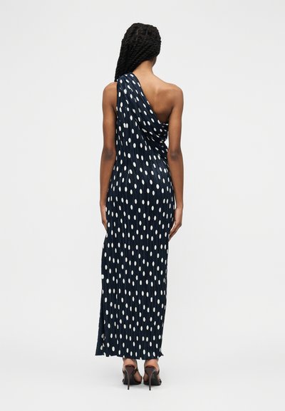 Γυναίκα με μακριά πλεξούδα που φοράει ένα μπλε σκούρο φόρεμα με πουά λευκές βούλες και μαύρα ψηλοτάκουνα σανδάλια, ορατή από πίσω.