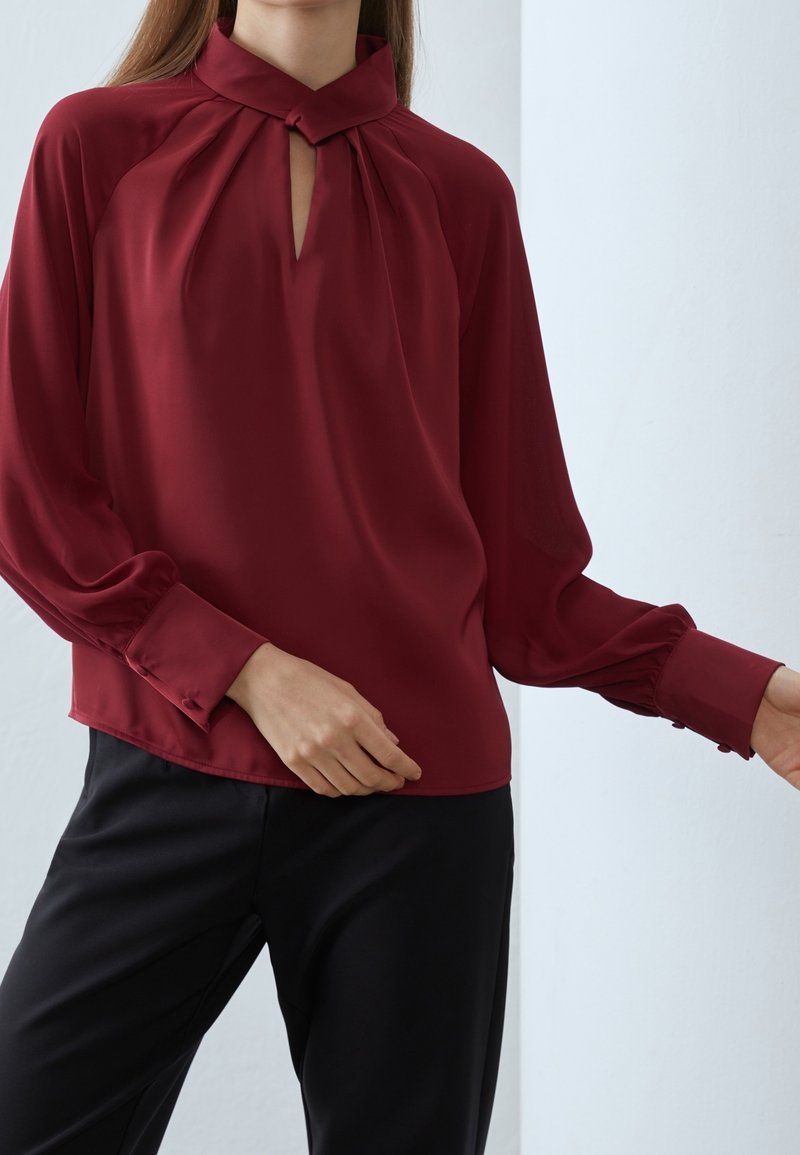 Blouse bordeaux avec un col montant, un détail en forme de trou de serrure et des manches longues. Texture lisse, accents plissés et poignets boutonnés. Associée à un pantalon noir.