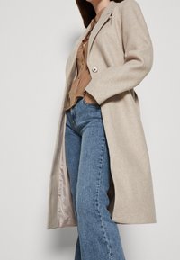 Béžový vlněný kabát smíchaný s jinými materiály, s propracovaným střihem, jedním knoflíkem a měkkou texturou, přeložený přes světle hnědou blůzu a modré džíny.