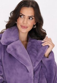 Cappotto in pelliccia sintetica viola con un grande colletto, tessuto morbido, maniche larghe e vestibilità rilassata. Accentuato con unghie naturali e orecchini a cerchio.