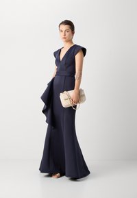 Robe bleu marine, longue jusqu'au sol, avec des manches courtes, décolleté en V profond et détail de volants asymétriques ; associée à un sac à main crème et des talons argentés.