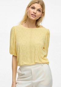 Blouse jaune à manches courtes avec un design en œillet texturé, col rond et manches bouffantes. Associée à un pantalon crème.