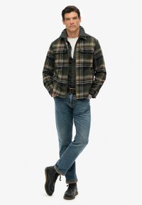 Camicia di flanella a quadri in verde, giallo e blu navy, indossata sopra una camicia a bottoni scura con jeans blu. Gli stivali neri con lacci completano l'outfit.