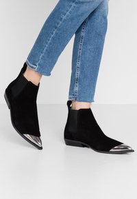 Botas de tobillo de ante negro con punta metálica puntiaguda, paneles laterales elásticos y un tacón bajo apilado, combinadas con jeans de mezclilla recortados.