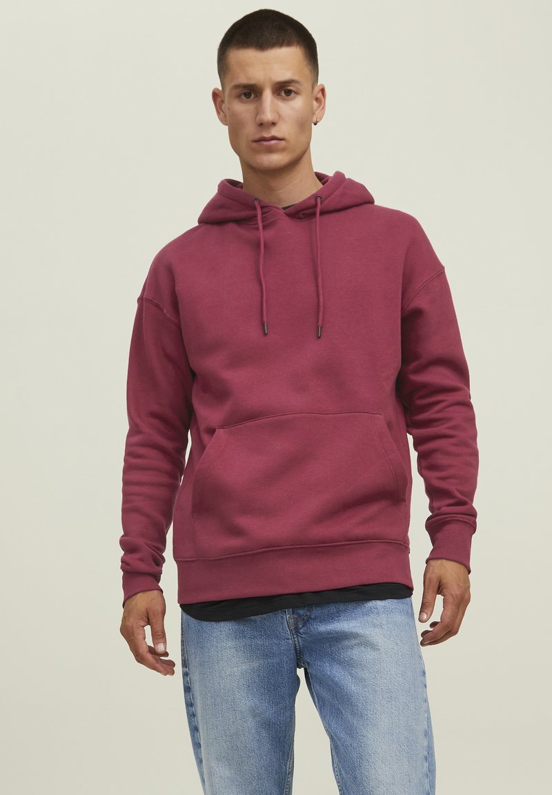 Hoodie bordeaux realizzato in tessuto morbido, dotato di tasca a marsupio, cordini e polsini a coste. Indossato con jeans di denim blu.