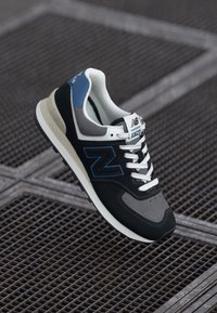 Black and grey sneaker with blue accents, featuring a textured surface, mesh tongue, and white laces. Model "574" displayed on the tongue.
