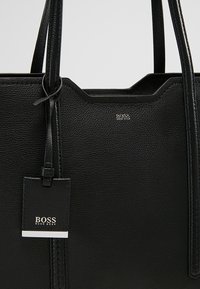 Sac cabas en cuir noir texturé avec doubles poignées et une étiquette suspendue embossée du logo « BOSS HUGO BOSS » en argent.