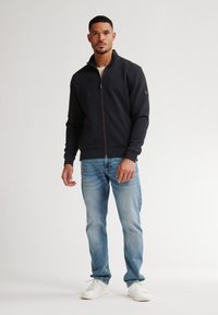 Giacca nera con zip e colletto alto, tessuto strutturato e una sottile patch con logo. Abbinata a jeans azzurri chiari e scarpe bianche.