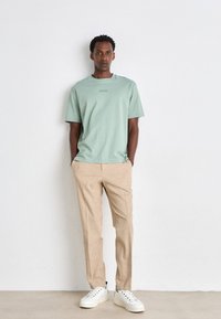 Lysegrønn bomull t-skjorte med en liten logo, kombinert med beige skreddersydde bukser og hvite sneakers, stående mot en lys bakgrunn.