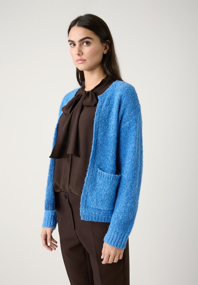 Blau strukturiertes Cardigan mit gerippten Bündchen und offener Vorderseite, mit einer aufgesetzten Tasche. Getragen über einer dunklen Bluse und braunen Hosen.