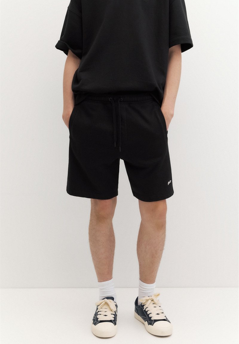 Personne portant un short noir avec cordon, un t-shirt noir, des chaussettes blanches et des baskets noires et blanches, debout sur un fond uni.