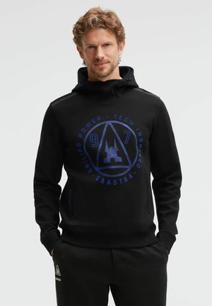 Man with light brown curly hair wearing a black hooded sweatshirt with blue logo text "Gaastra 97 Sailing Power Tech Inspired."