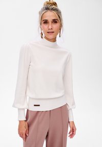 Biała bluzka z długim rękawem, wysoka stójka i prążkowane mankiety. Miękki dzianinowy materiał z dopasowanym dołem i małym logo z przodu.