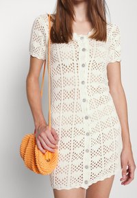 Abito di maglia color panna con maniche corte, chiusura con bottoni e motivo simile a pizzo. Porta una borsa a tracolla rotonda arancione intrecciata.