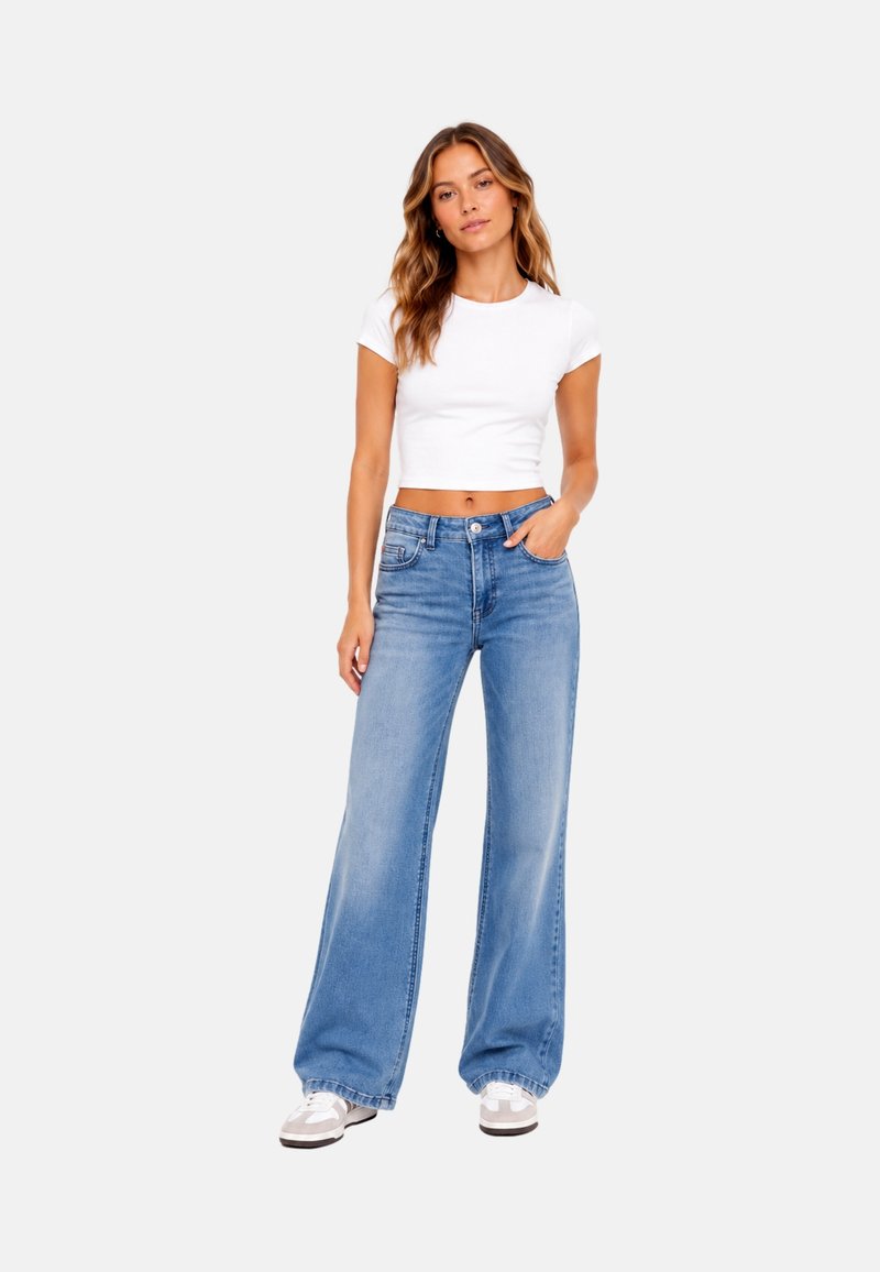 Femme portant un t-shirt court blanc, un jean bleu taille haute à jambes larges et des baskets blanches, debout avec une main dans la poche.