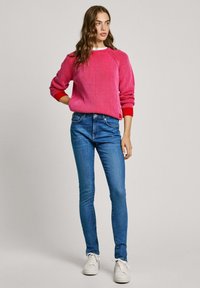 Růžový žebrovaný svetr s červenými akcenty, laděný k modrým těsným džínům a bílým teniskám. Modelka stojí na neutrálním pozadí.