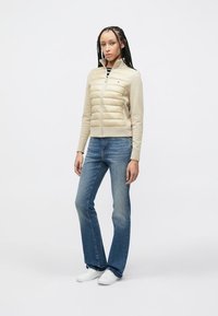 Μπεζ πανωφόρι με επένδυση, ψηλό γιακά και κλείσιμο με φερμουάρ, συνδυασμένο με καμπάνα μπλε τζιν και λευκά αθλητικά παπούτσια. Λείο ύφασμα και καθαρός σχεδιασμός.