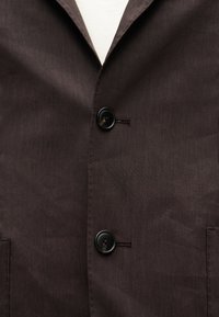 Μαύρα κουμπιά σε σκούρο καφέ σακάκι πάνω από λευκό πουκάμισο, που δείχνουν το εμπρόσθιο κλείσιμο και την υφή του υφάσματος σε κοντινή όψη.