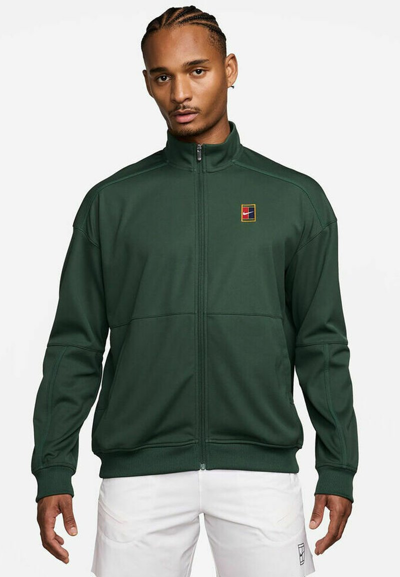 Giacca verde con zip realizzata in tessuto morbido, caratterizzata da un piccolo logo ricamato in alto a sinistra e cuciture a contrasto lungo le spalle.