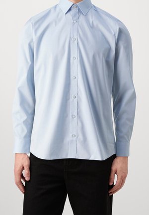 Man die een lichtblauw overhemd met lange mouwen en een puntige kraag draagt, gecombineerd met een zwarte broek, staand tegen een effen achtergrond.