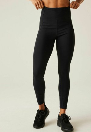 Athletische Person trägt schwarze Leggings mit hohem Bund und schwarze Laufschuhe und steht vor einem schlichten hellen Hintergrund.