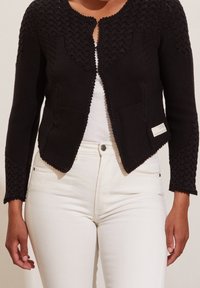 Černý pletený kardigan s texturovaným vzorem stehů, kulatým výstřihem, předním otevřením a přiléhavými rukávy. Kombinován s bílými kalhotami.