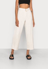 Szerokie spodnie w jasnobeżowym lnie, z wysokim stanem, fałdami z przodu i złotą klamrą paska w kształcie pierścienia. Połączone z czarnymi klapkami.