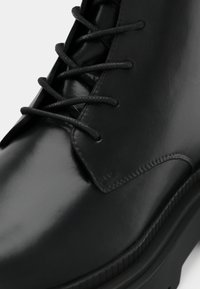 Black leather ankle boot with a glossy finish, round toe, six eyelets, black laces, and a textured rubber sole.