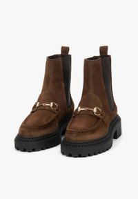 Brązowe zamszowe botki za kostkę z masywną czarną gumową podeszwą, elastycznymi bocznymi panelami i złotym detalem na przodzie.