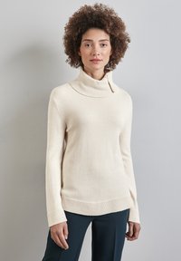 Maglione a collo alto color crema chiaro con colletto e polsini a coste, texture liscia e linea dritta, indossato sopra pantaloni scuri.