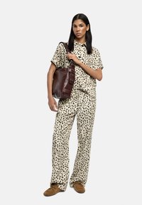 Kobieta w beżowych i czarnych, w kropki, luźnych spodniach oraz koszulce z krótkim rękawem, trzymająca brązową skórzaną torbę na ramię, stojąca na białym tle.