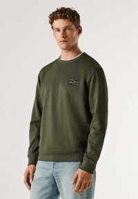 Alyvuogių žalia džemperis su apvalia iškirpte, ilgomis rankovėmis, ribinėmis rankogaliais ir stačiakampiu lipduku ant krūtinės su prekės ženklo logotipu.