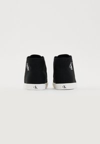 Une paire de baskets montantes noires avec des semelles blanches et le logo "cK" sur le talon et le côté, présentée de dos sur un fond blanc uni.