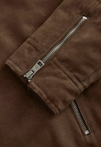 Brūna zamša jaka ar piedurkni, kurā ir metāla rāvējslēdzējs ar taisnstūra vilkšanas cilpu, šūtas malas un mīksta, matēta tekstūra.