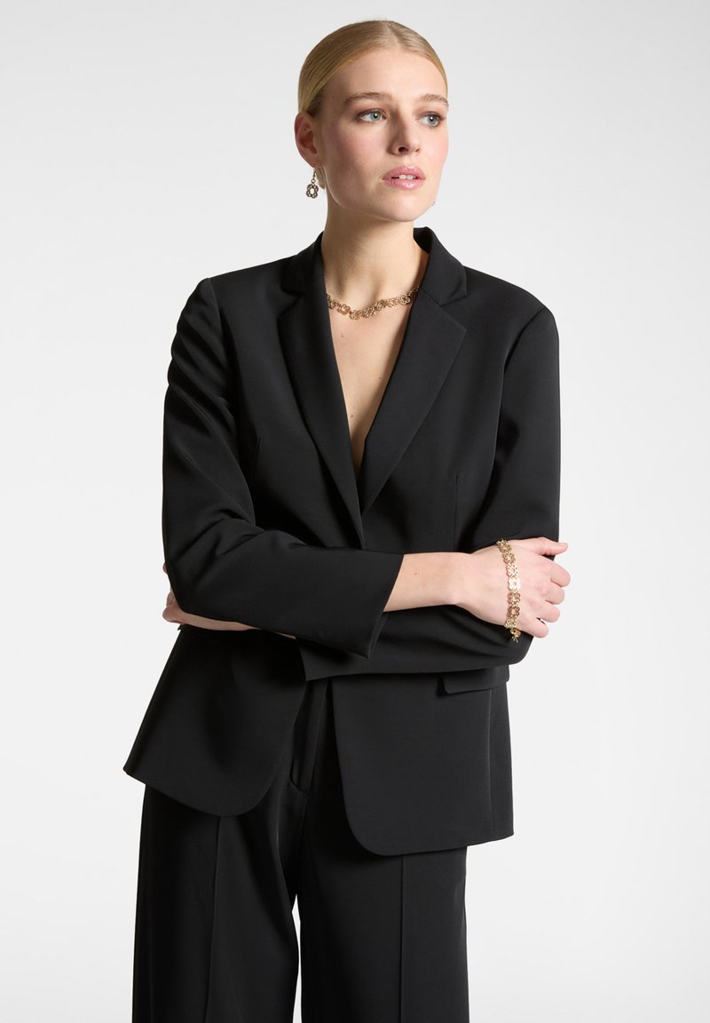 Blazer nero su misura con rever a punta e maniche lunghe, abbinato a pantaloni a vita alta coordinati. Modella che indossa gioielli in oro.