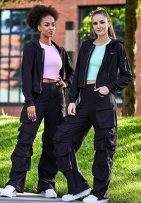 Dva modely nosia zladené outfity, ktoré pozostávajú z čiernych cargo nohavíc s viacerými vreckami. Jeden model má na sebe ružový crop top, zatiaľ čo druhý má mätovo zelený top.