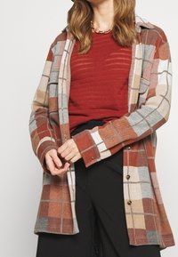 Giacca a camicia a quadri nei toni ruggine, beige e grigi, con chiusura a bottoni e vestibilità rilassata. Tessuto morbido e strutturato con maniche lunghe.