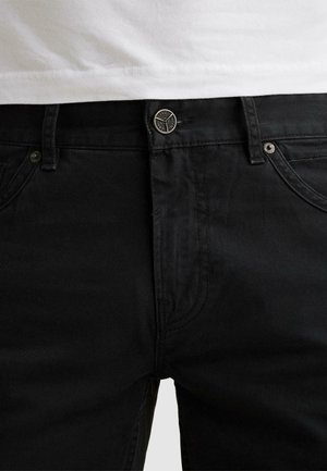 Detail černých džínů z denimu v oblasti pasu s kovovým knoflíkem a poutky na pásek, doplněných bílou košilí zastrčenou dovnitř.