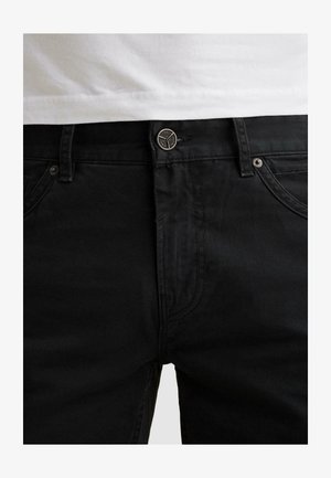 Detail černých džínů z denimu v oblasti pasu s kovovým knoflíkem a poutky na pásek, doplněných bílou košilí zastrčenou dovnitř.