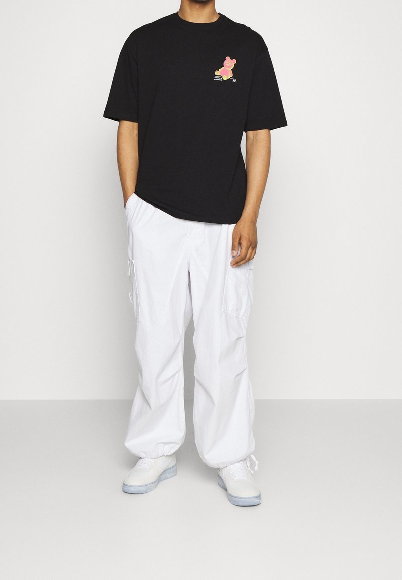 Mustahiilaa musta t-paita, jossa on sarjakuvamainen karhukuva rinnassa, yhdistettynä löysiin valkoisiin cargo-housuihin ja valkoisiin lenkkareihin.