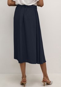 Fustă midi plisată de culoare bleumarin cu talie elastică, design fluid și textură netedă, purtată cu pantofi cu toc transparent.