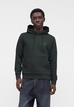Mężczyzna stoi, ubrany w ciemnozieloną bluzę z kapturem z logo "Jack & Jones" i niebieskie dżinsy, patrzy do przodu na jasnym, jednolitym tle.