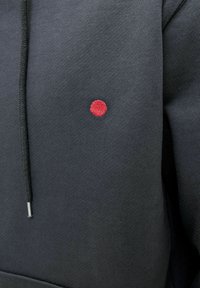 Čierna mikina s kapucňou z texturovanej látky, so malým červeným vyšitým kruhom na hrudi a rovnými šedými šnúrkami so striebornými agletmi.