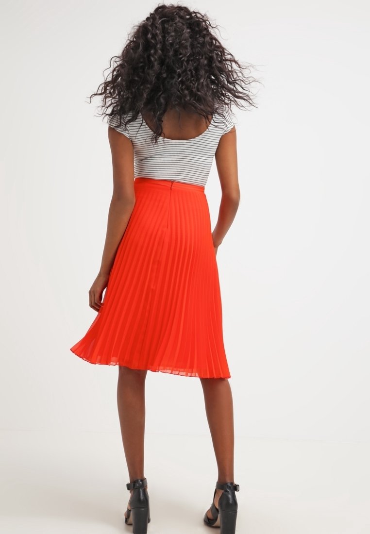 Röd plisserad kjol i lätt tyg, knälång med slät midja, parat med en randig topp och svarta klacksandaler.