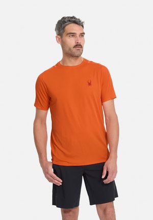 Homme aux cheveux gris courts et à la moustache portant une chemise orange à manches courtes avec un petit logo d’araignée et un short noir, debout et regardant vers la droite.