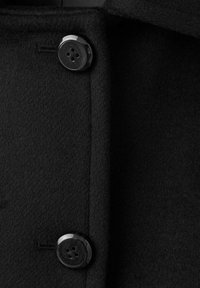 Abrigo de lana negra que muestra un primer plano de la textura del tejido y dos botones redondos negros con cuatro agujeros cosidos en la costura frontal.