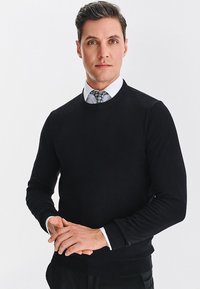 Czarny sweter dzianinowy z okrągłym dekoltem i prążkowanymi mankietami, noszony na białej koszuli z wzorzystym krawatem. Model stoi z rękami złożonymi.