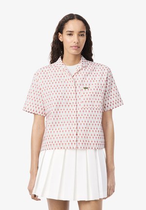 Vrouw die een wit overhemd met korte mouwen draagt met een klein rood patroon en een witte geplooide rok, staand tegen een effen achtergrond.