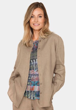 Beige Leinenjacke mit Knopfverschluss, kombiniert mit einem farbenfrohen, gemusterten Hemd, das geometrische Formen und lebhafte Farben zeigt.