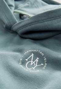 Bluza z kapturem w jasnym odcieniu teal, wykonana z miękkiego materiału. Posiada biały haftowany logo z napisem "MANHATTAN ISLAND" oraz "BIG SMILE CLUB."
